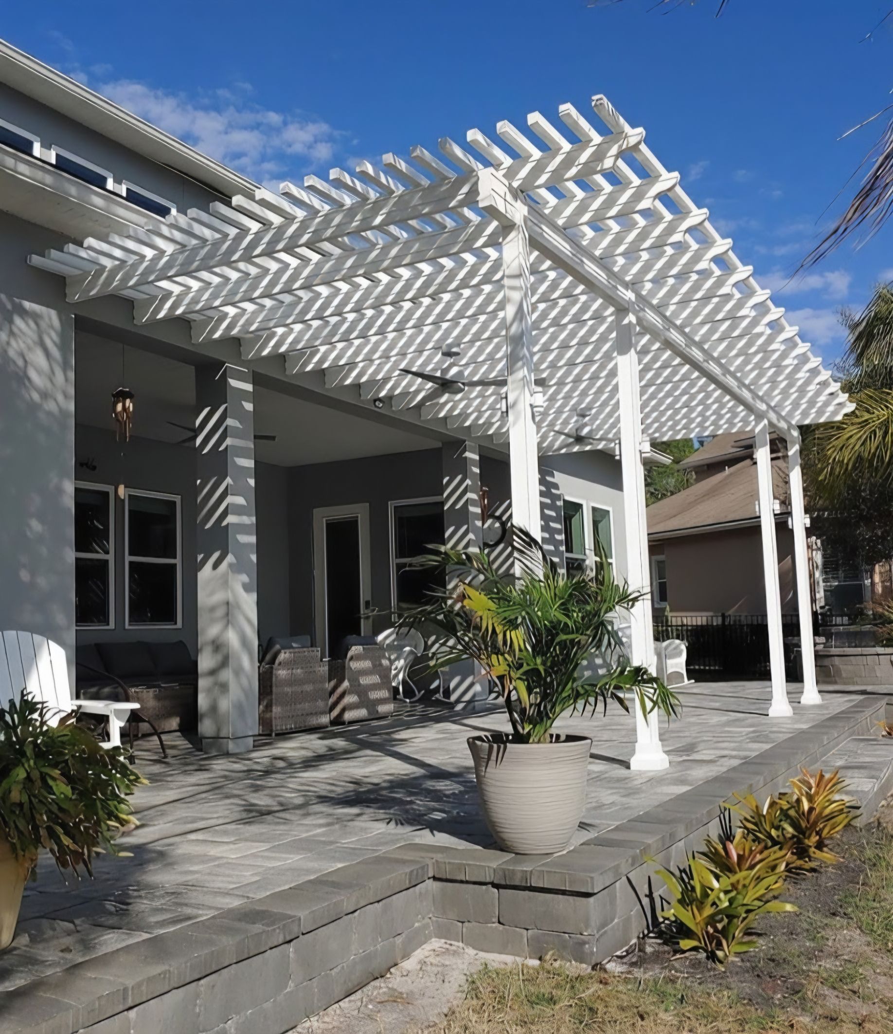 A white pergola extends from a gray house over a paved patio, featuring support columns and potted plants.