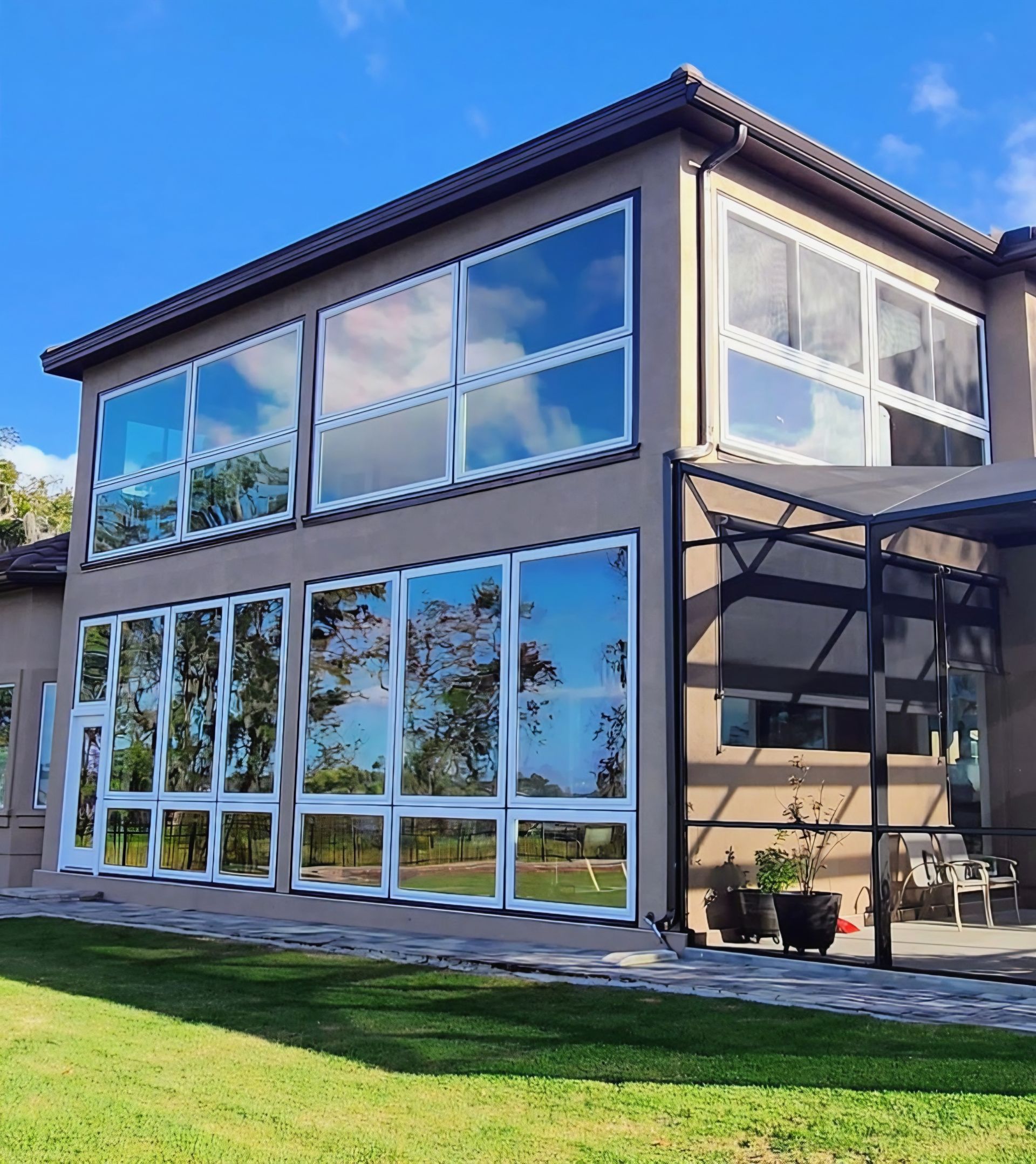 A two-story tan house with many large, reflective windows, a stone patio, and a grassy lawn under a clear blue sky.