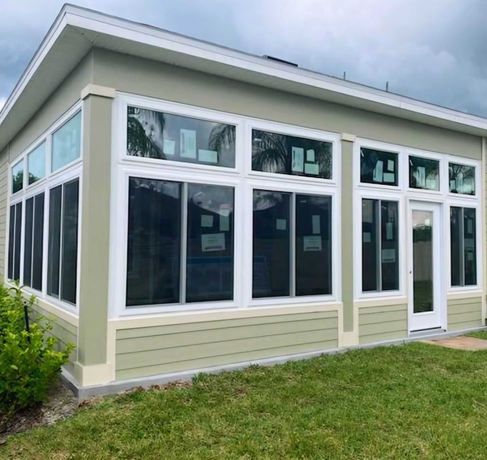 A light green, modern sunroom addition with large white-framed windows, a glass door, and horizontal siding on a grassy lawn.