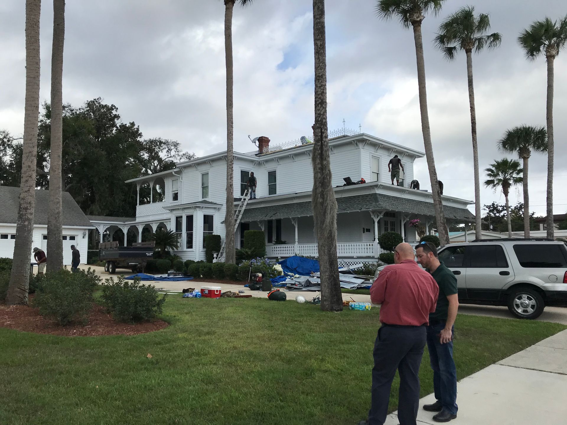 Two people stand on a lawn observing workers on the roof of a large, two-story white house surrounded by palm trees.