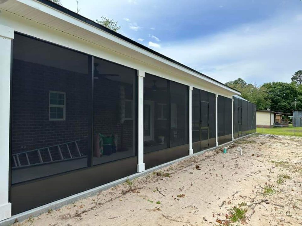 Screened-in porch on a house with a white trim and black screens, set on sandy ground with a cloudy sky.
