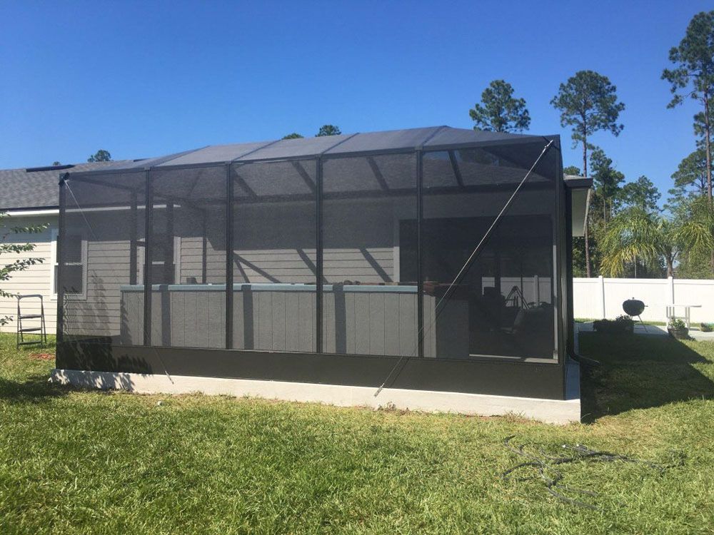 Screened-in patio enclosure with a hot tub inside, black screen and frame, set on a concrete foundation, in a grassy backyard under a blue sky.