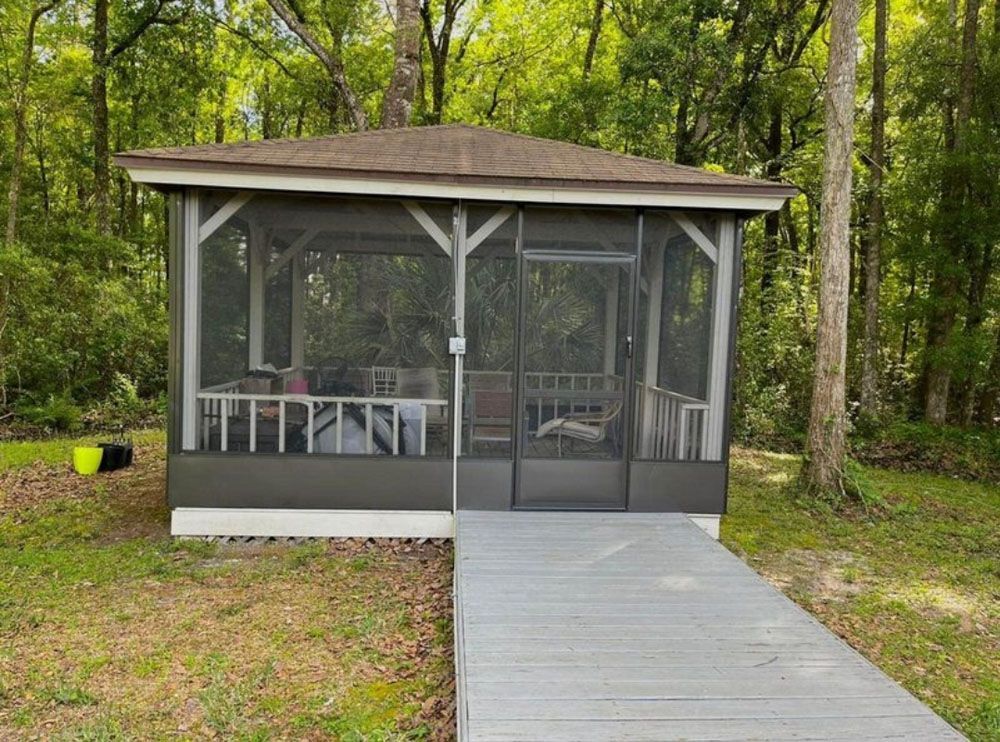 Screened gazebo with gray deck, in a wooded area. Brown roof, white trim.