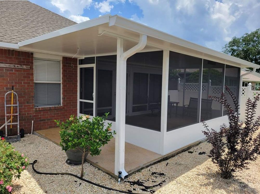 Screened-in patio extension attached to a brick house. White frame, black screens, concrete floor, gravel yard.