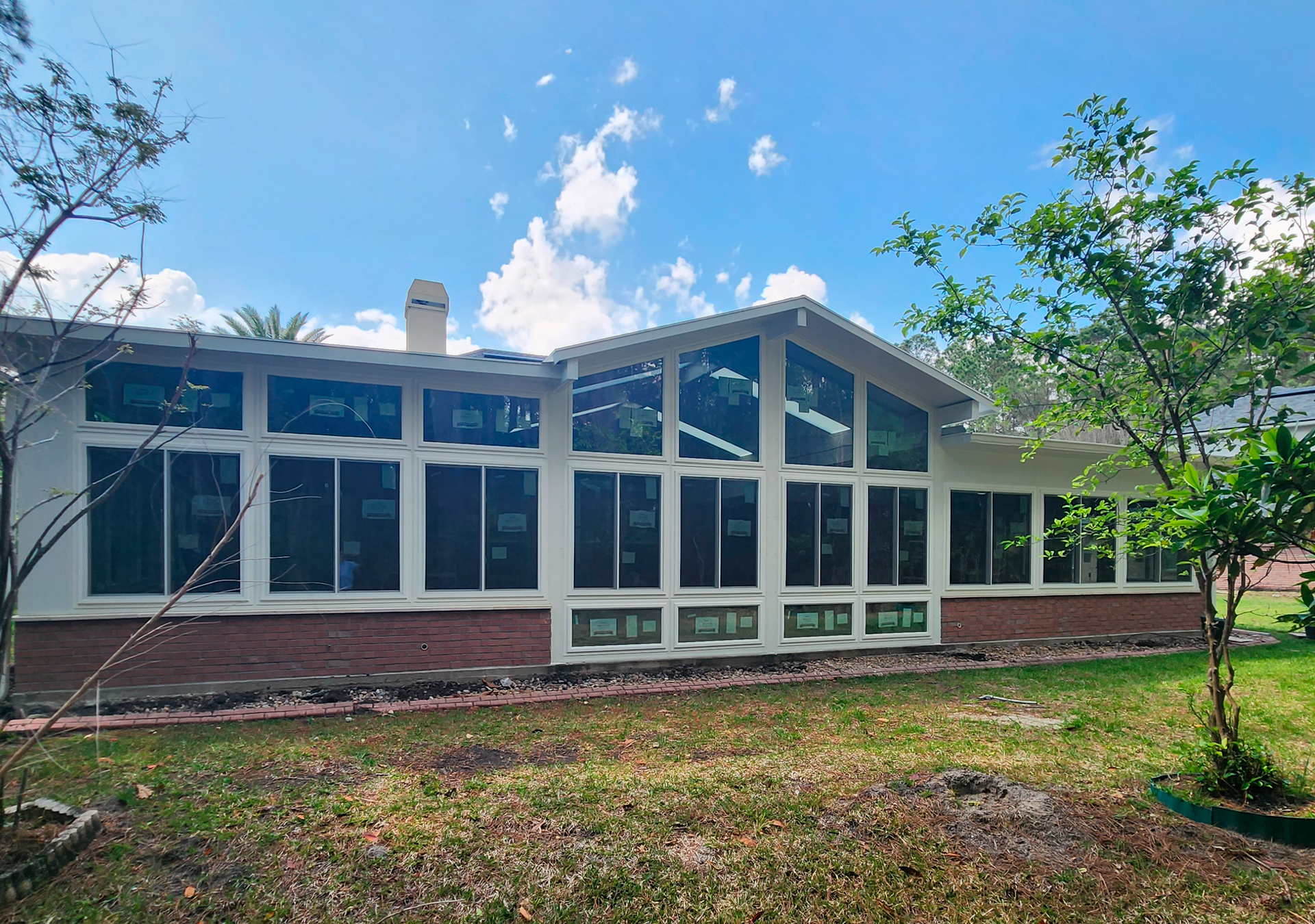 Backyard view of a white-walled house with many windows, brick foundation, and trees against a blue sky.