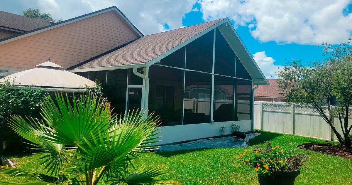 Screened patio attached to a house with a tile roof, surrounded by lawn and shrubs; blue sky.