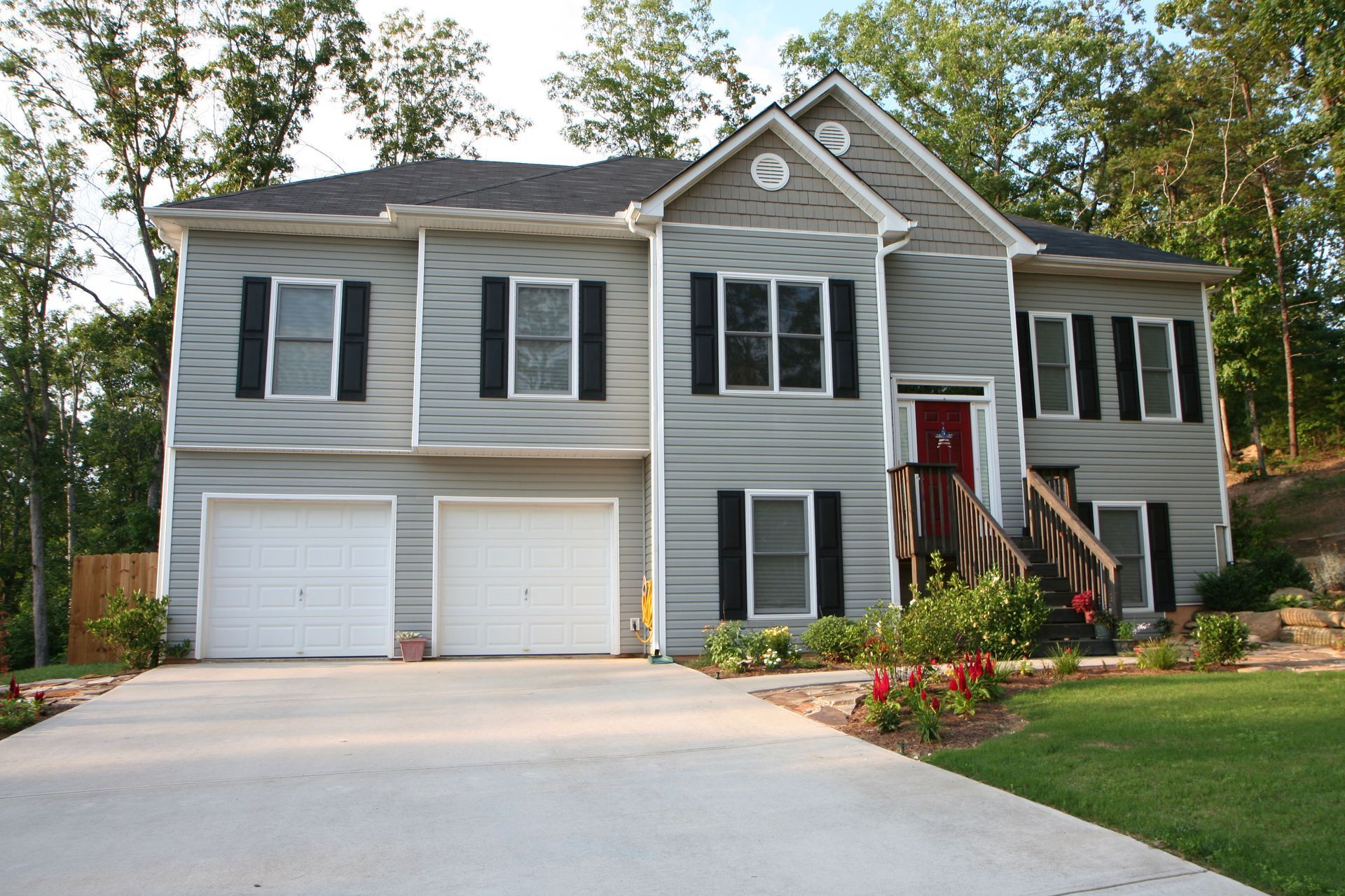 Two-story gray house with white garage doors and a red front door, surrounded by trees and a concrete driveway.