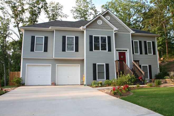 Two-story gray house with white garage doors and a red front door, surrounded by trees and a concrete driveway.