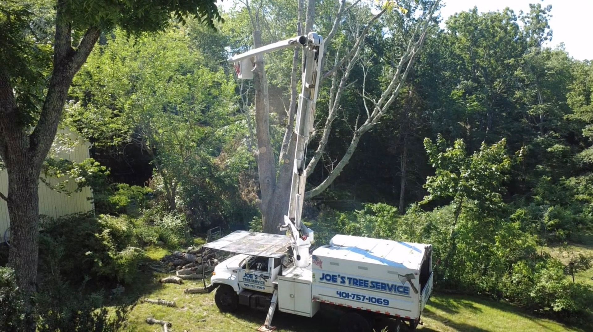White utility truck with a raised boom lift in a wooded area beside dense green trees.