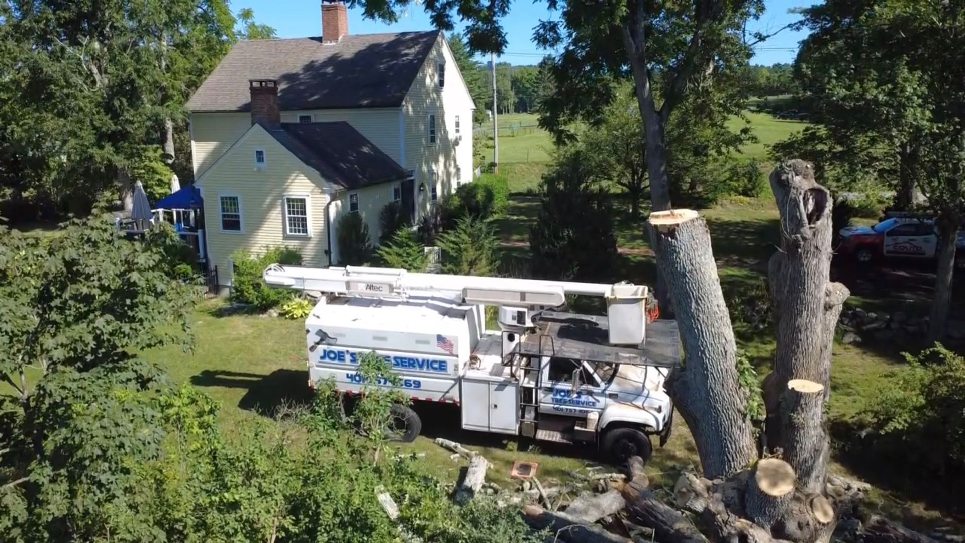 Bucket truck working beside a white house with cut tree trunks and branches in a sunny yard