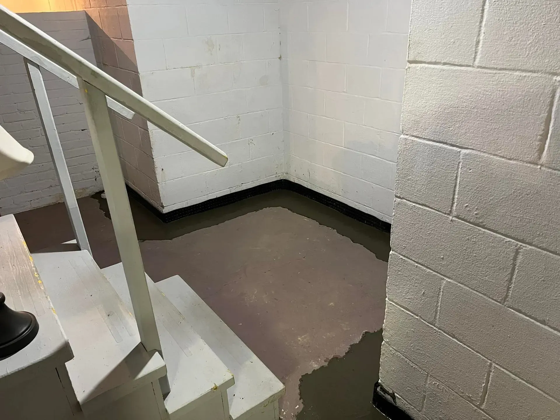 Small stair landing with white cinderblock walls, white railing, and a brown floor with a worn patch.
