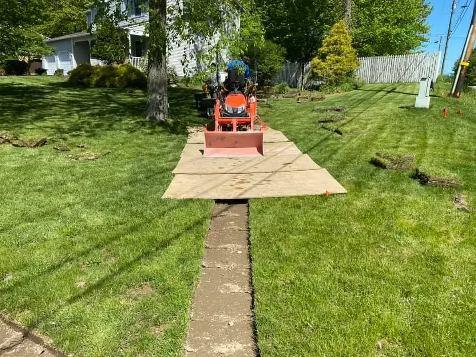 Orange lawn mower on plywood over a grassy yard, trimming along a narrow path between trees.