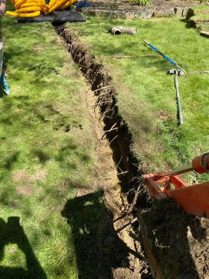 A deep trench dug through a grassy yard, with tools and a wheelbarrow nearby.