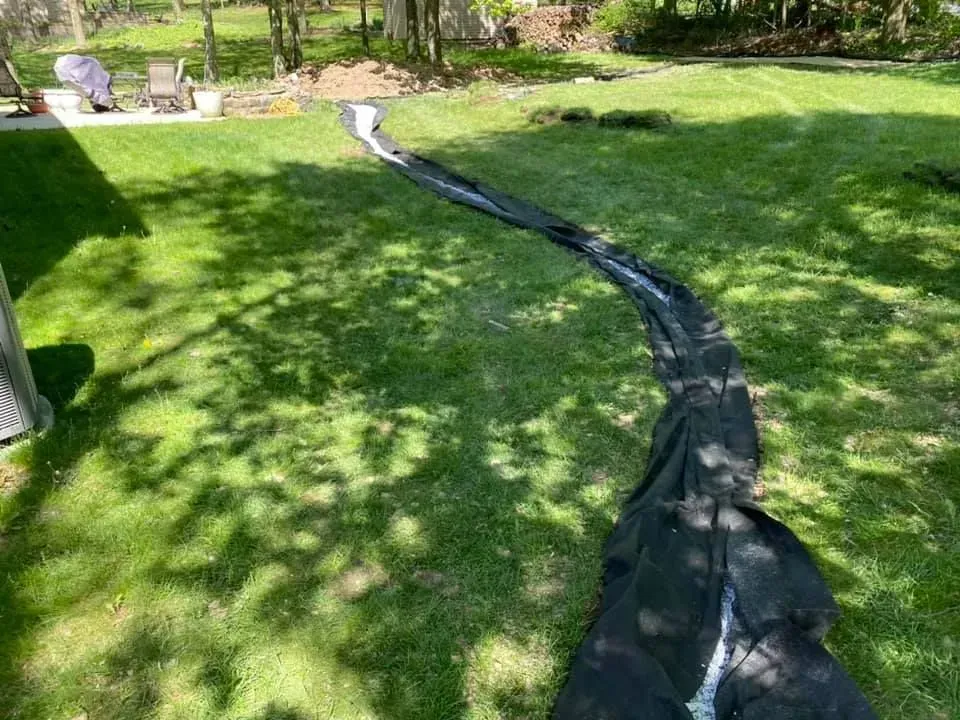Black drainage hose across a green lawn leading to a patio with chairs and trees