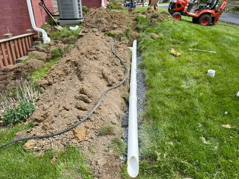 Trench beside a house with a white pipe laid in the ground and a small orange tractor in the distance.