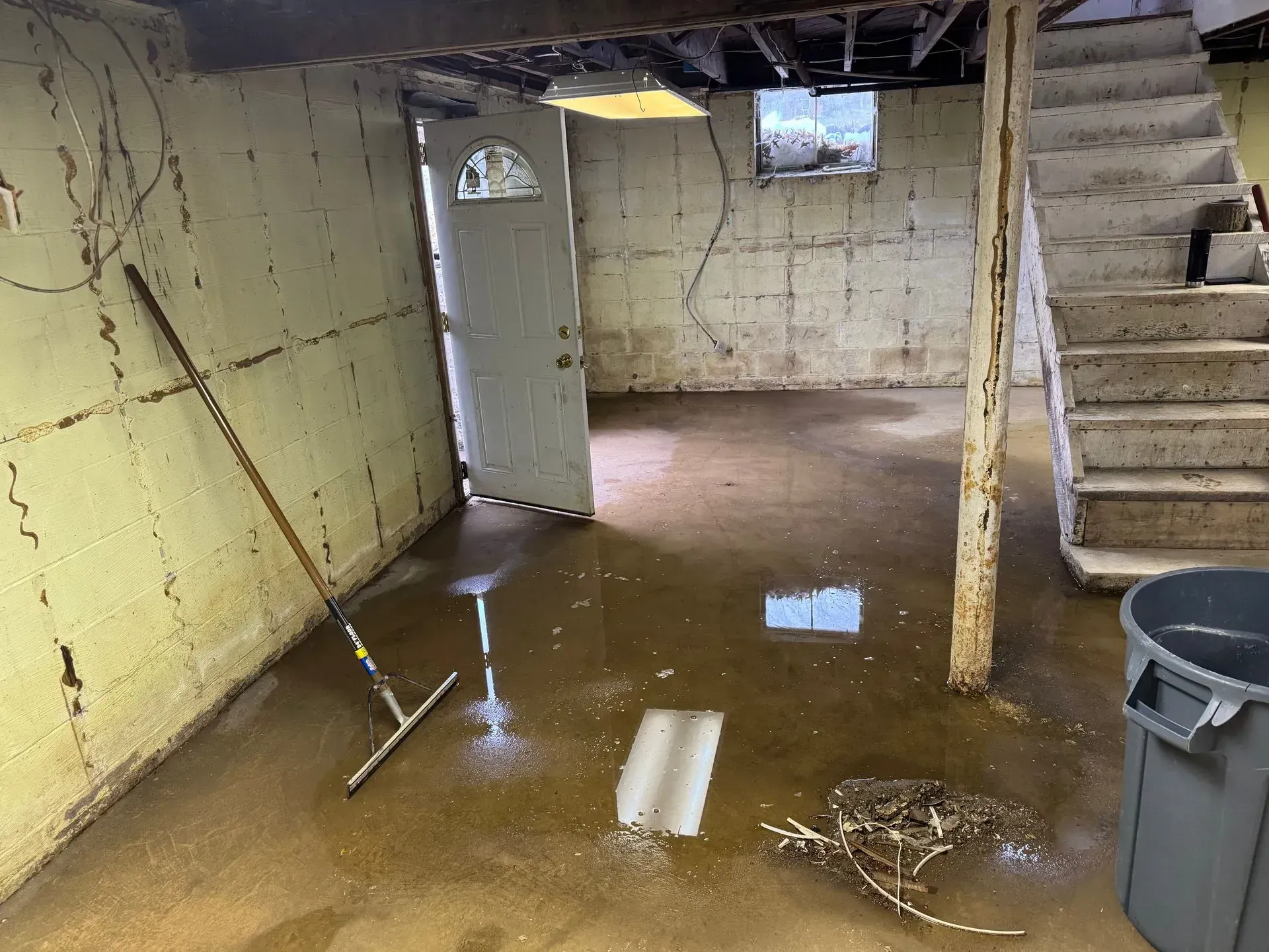 Flooded basement with muddy water, exposed insulation, open door, and stairs along the right wall.