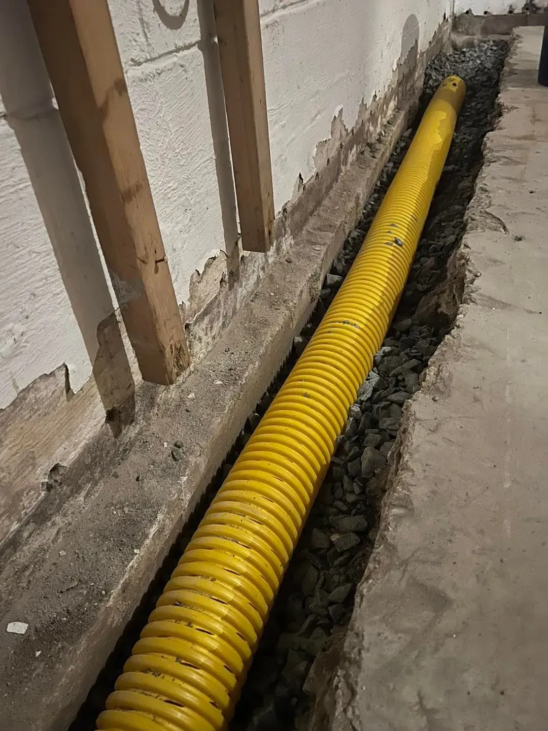 Yellow corrugated pipe laid in a trench along an unfinished wall inside a building.