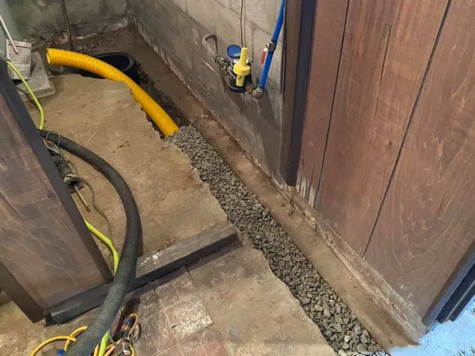 Trench with exposed pipes and gravel beside a wooden wall, with yellow and black hoses in a work area