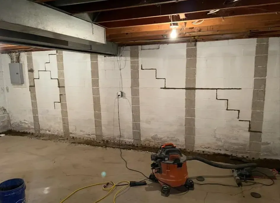 Basement wall with crack repairs, exposed ceiling beams, and a shop vac on the floor