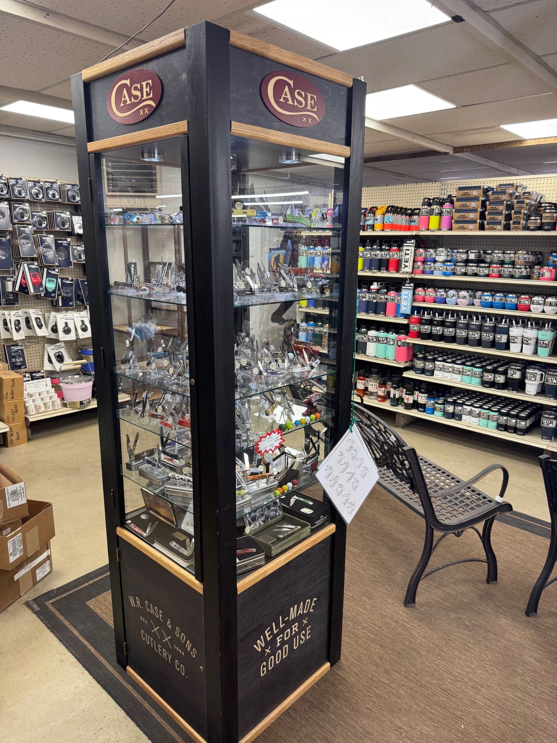 A tall, black Case-branded display case filled with knives in a retail store, next to shelves of merchandise and a bench.