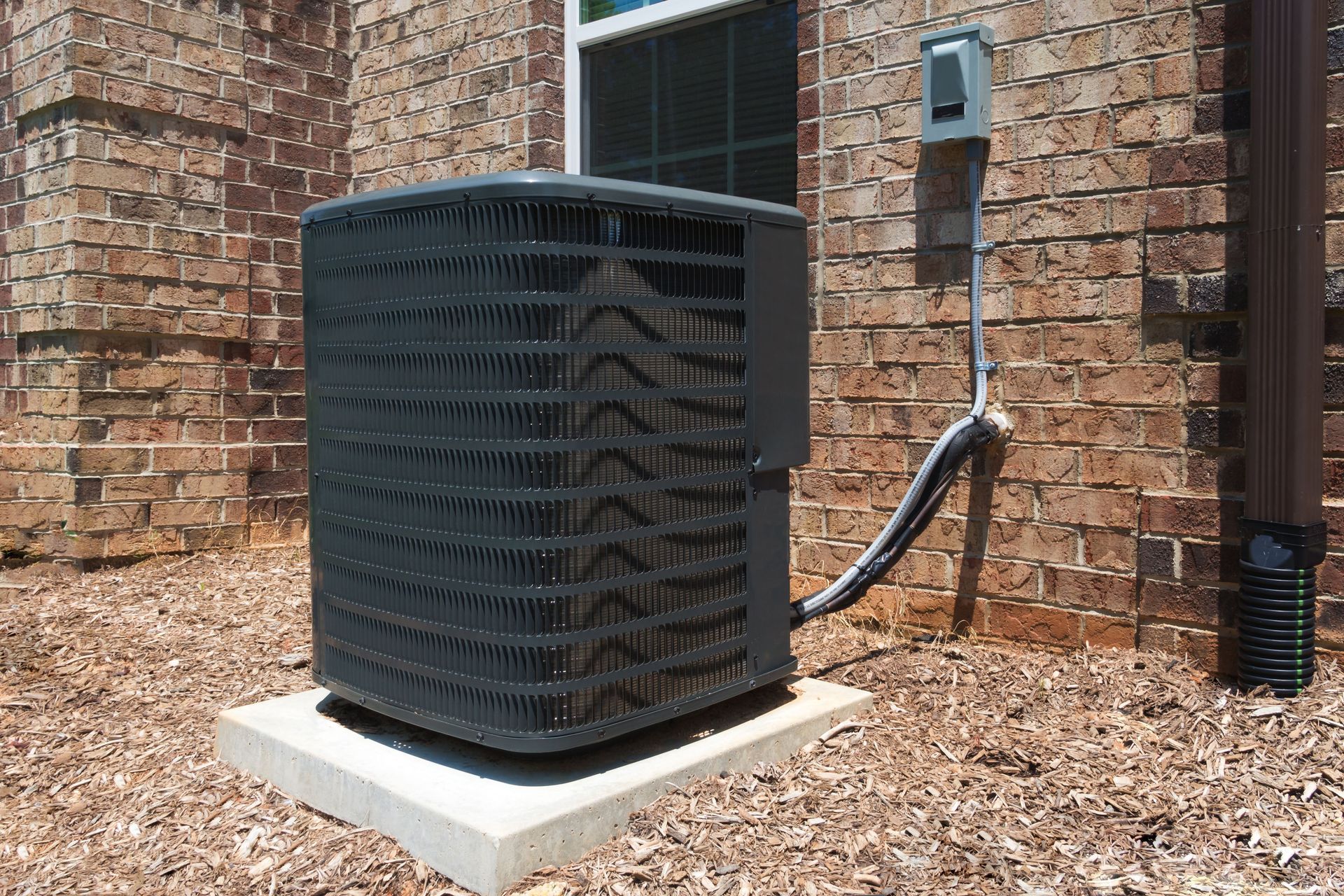 Outdoor air conditioning unit beside a brick house, with a black hose and utility box nearby.