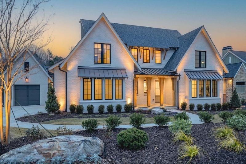 White farmhouse-style home at dusk, with lit windows, landscaping, and a garage.