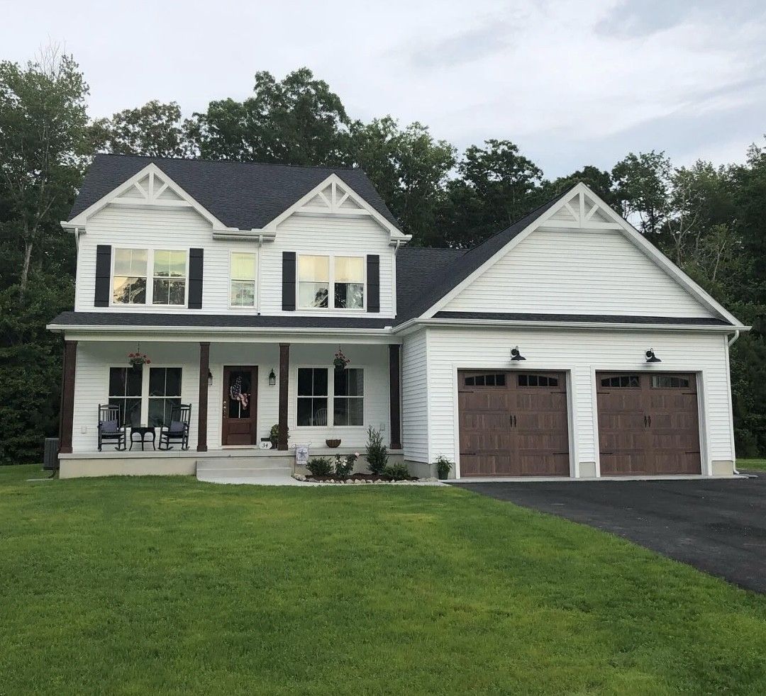 White two-story house with dark roof and brown garage doors, set in a green yard with trees in the background.