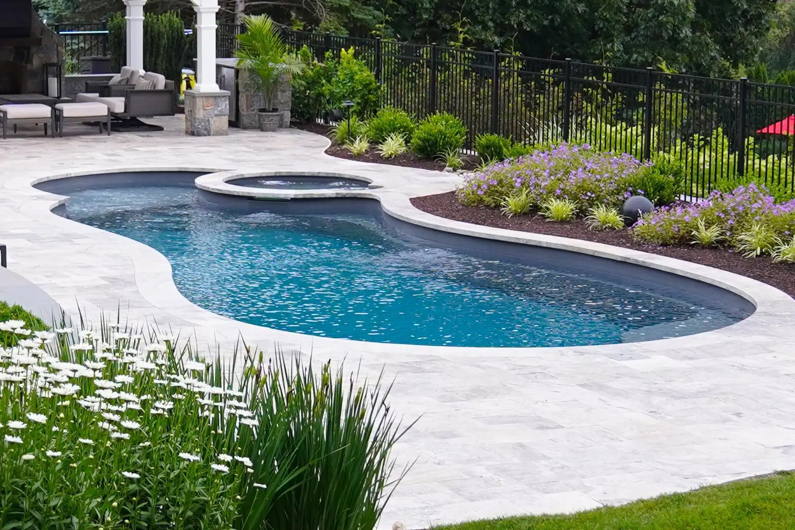 Pool with connected hot tub surrounded by light-colored stone patio and landscaped garden with flowers and greenery.