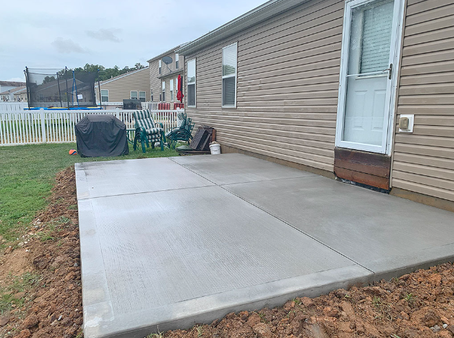 Newly poured concrete patio next to a beige house, with grass and a fence in the background.