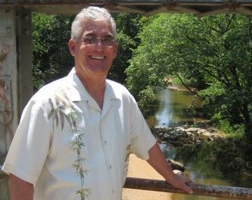 The business owner wearing a white shirt is standing on a bridge overlooking a river.