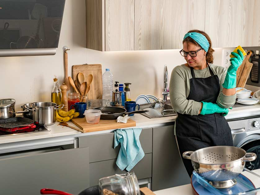 Woman in apron and gloves looks at a messy kitchen filled with dirty dishes and cooking utensils.