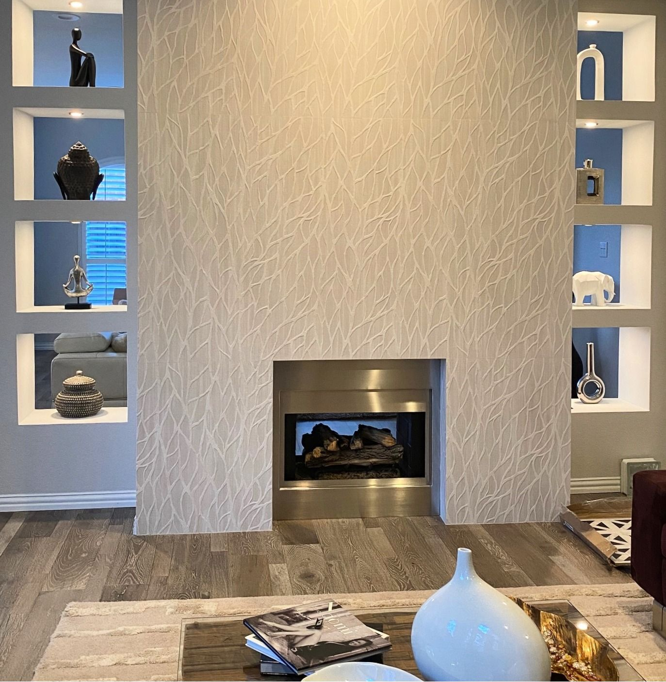Fireplace with built-in shelves displaying art objects. Neutral-toned textured wall, stainless steel fireplace, hardwood floor.
