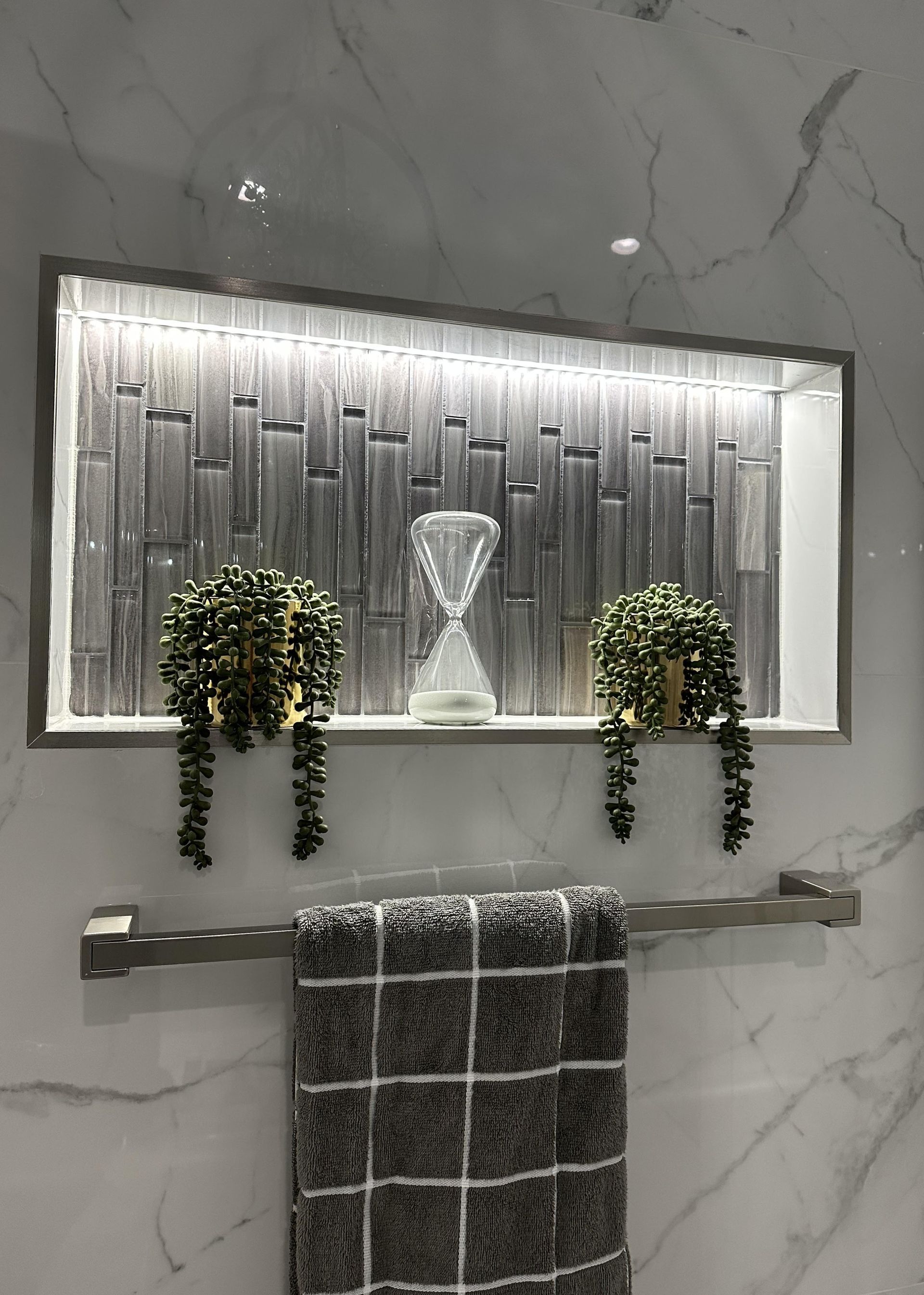 Bathroom niche with decorative gray mosaic tile, hourglass, trailing plants, and towel on bar.