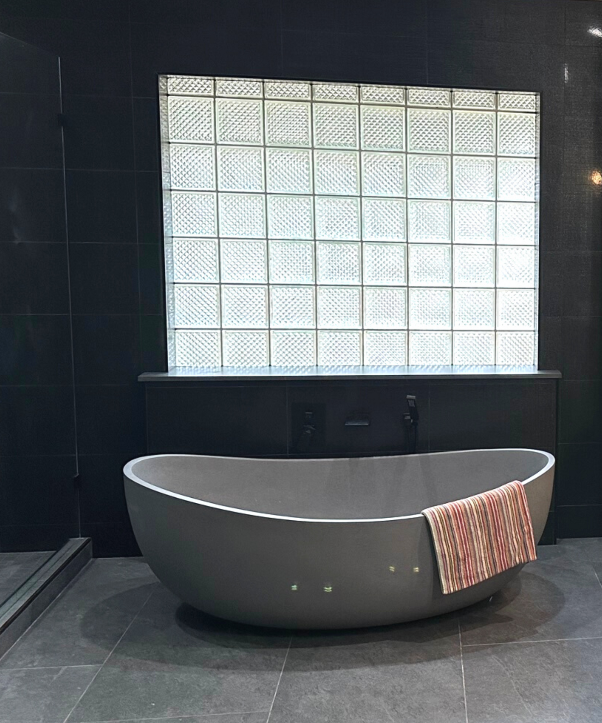 Oval gray bathtub in dark bathroom, glass block window above. Towel on tub edge.