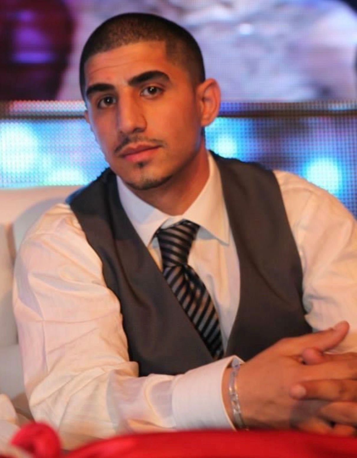 Man in white shirt and gray vest seated at a table, looking at the camera with a colorful blurred background