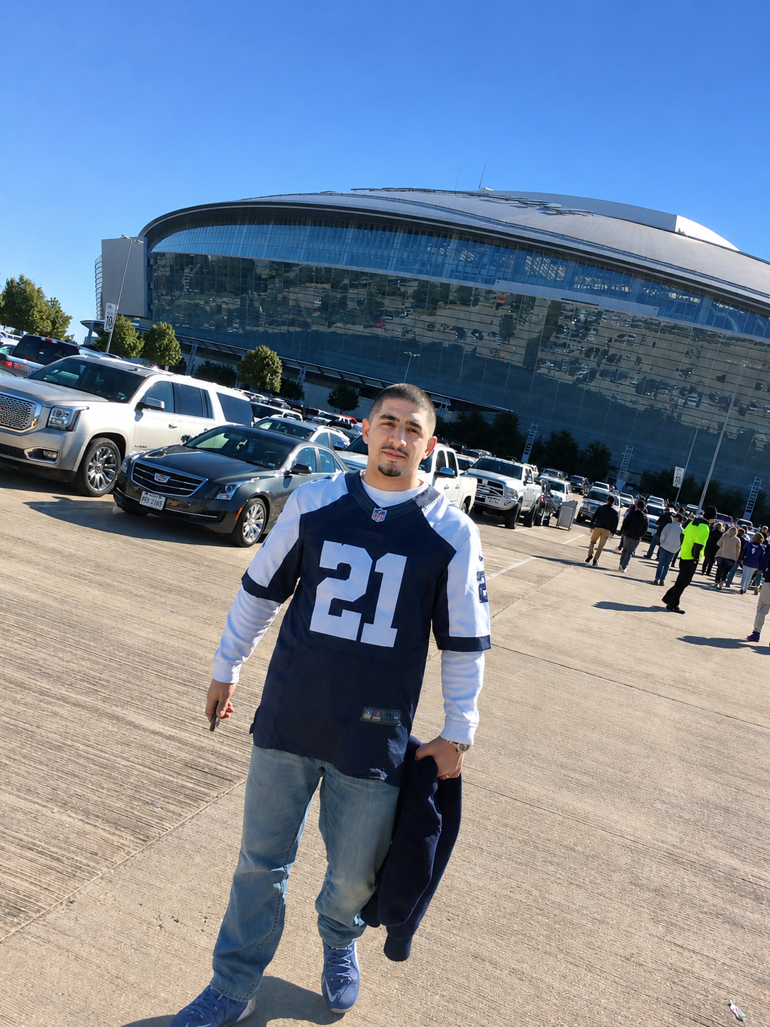 Man standing in a Cowboys jersey outside a large stadium on a sunny day, with parked cars nearby.