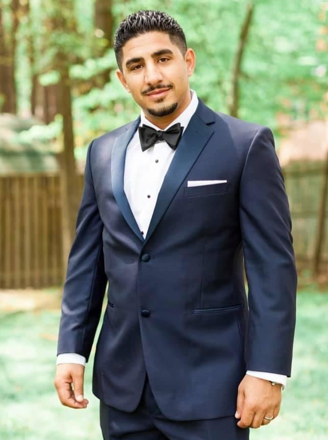 Man in a navy suit and bow tie standing outdoors with a blurred green background