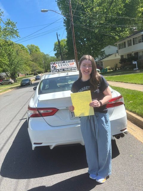 Person holding a yellow paper stands behind a white car on a sunny residential street.