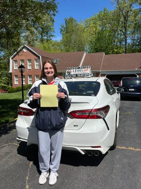 Woman standing behind a white sedan, holding a yellow sign in a driveway with houses and trees.