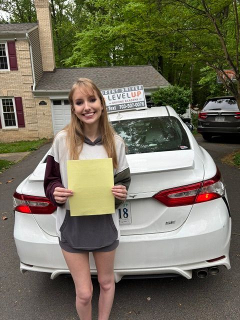 Woman holding a yellow paper in front of a white car in a driveway, with a house and trees behind.