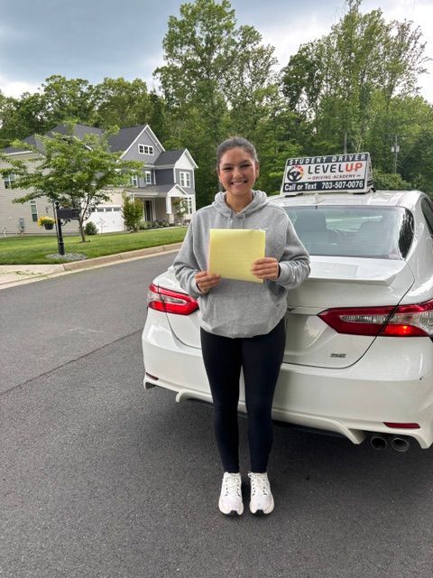Woman holding a yellow paper stands behind a white car on a suburban street with a “Level Up” sign in the background