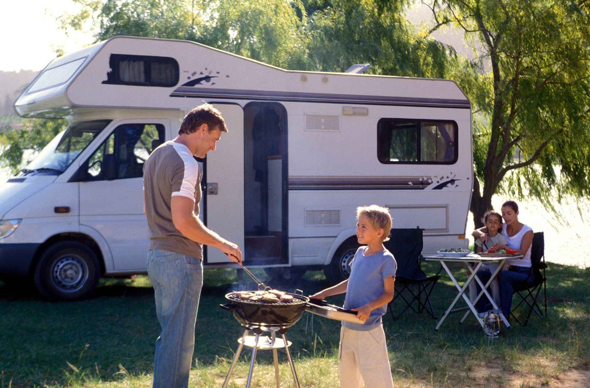 Family camping with a motorhome. Father grilling with son, mother and child seated at a table.