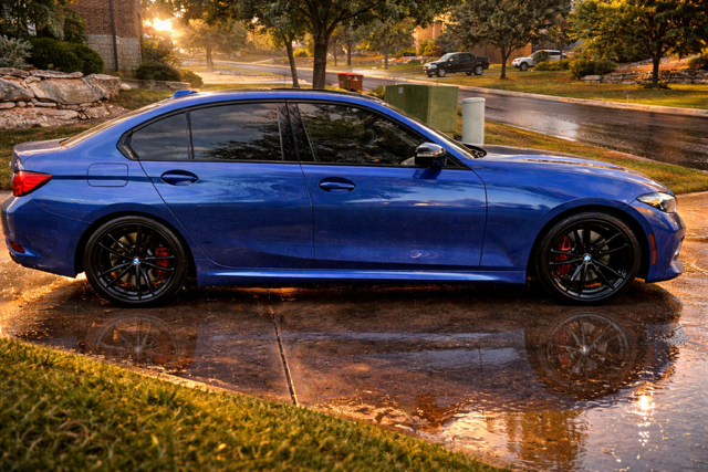 Blue sedan parked on a wet driveway at sunset, with trees and reflections in the background.