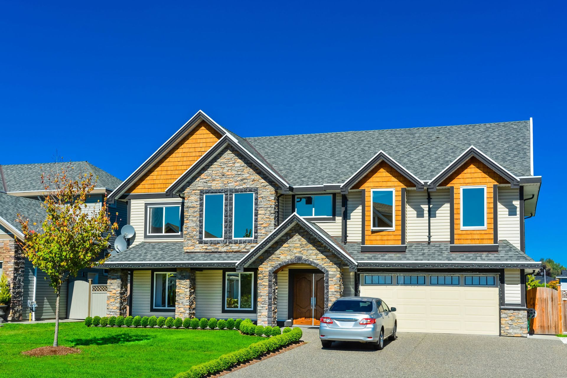 Two-story house with stone and wood accents, gray roof, blue sky, car in driveway, green lawn.