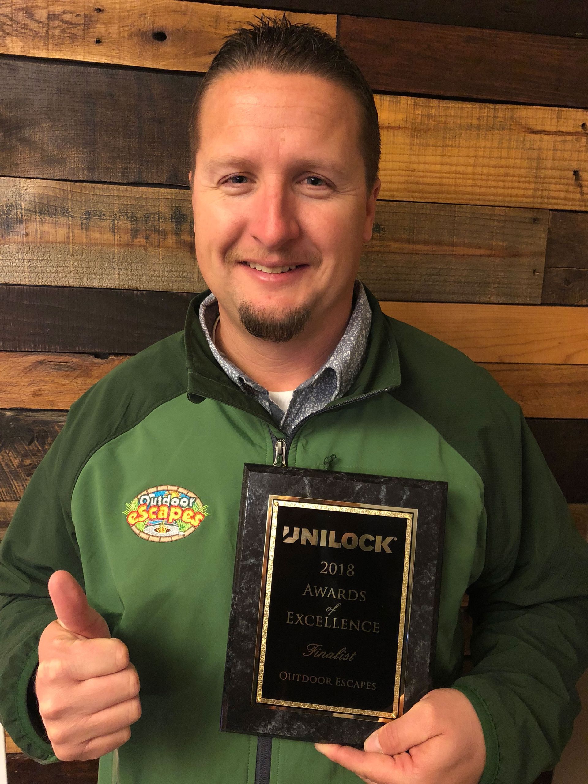 A smiling person in a green jacket holds a plaque and gives a thumbs-up against a wood-paneled wall.