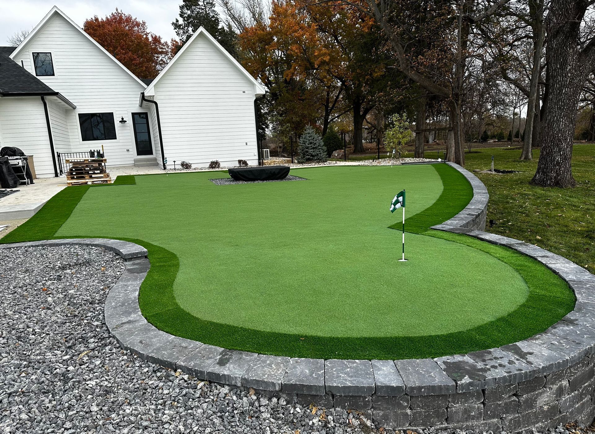 Backyard putting green with artificial turf, stone border, and a small flag beside a white house