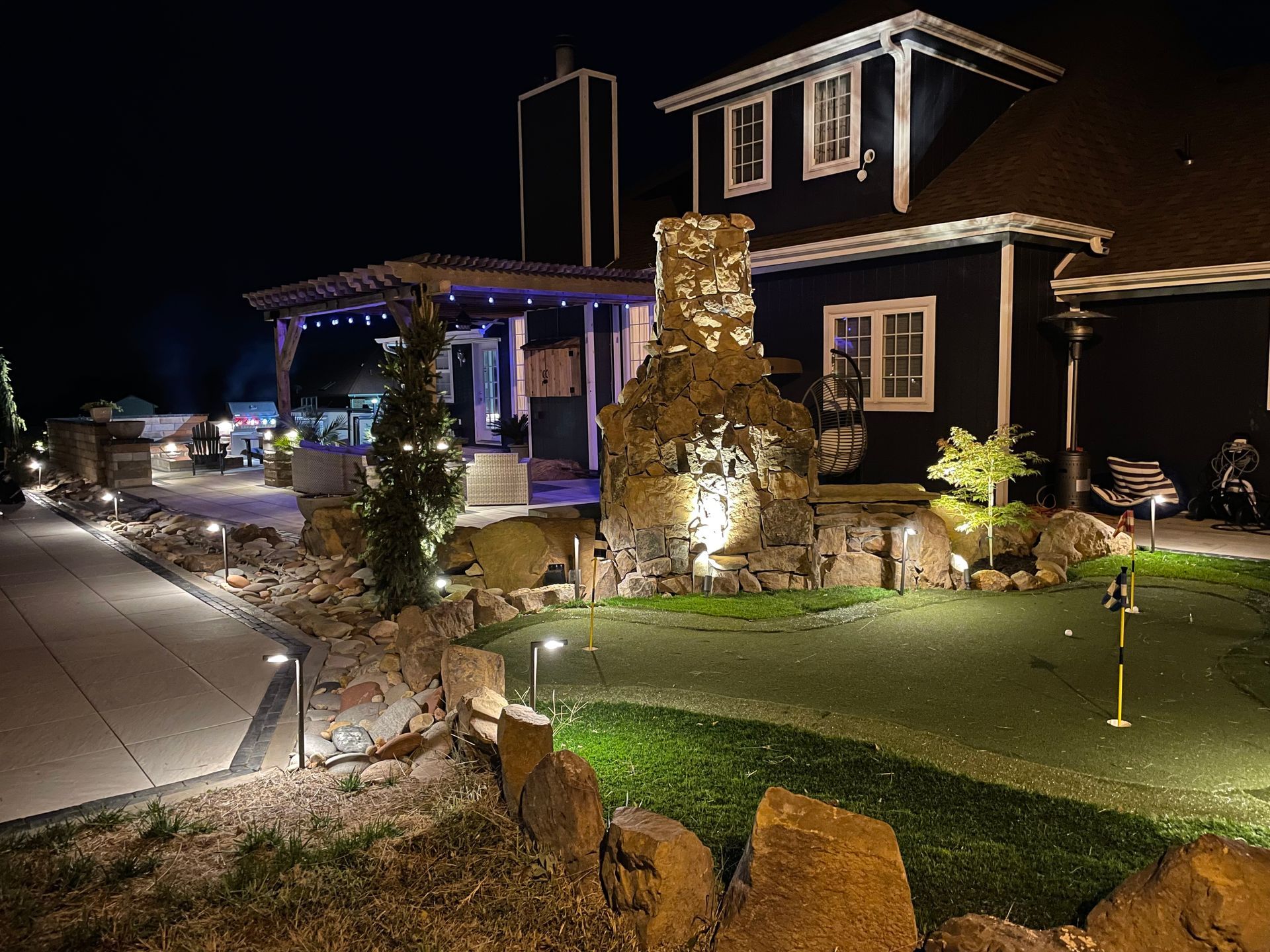 Lit house and front yard at night with walkway lights, stone chimney, and decorated landscaping.