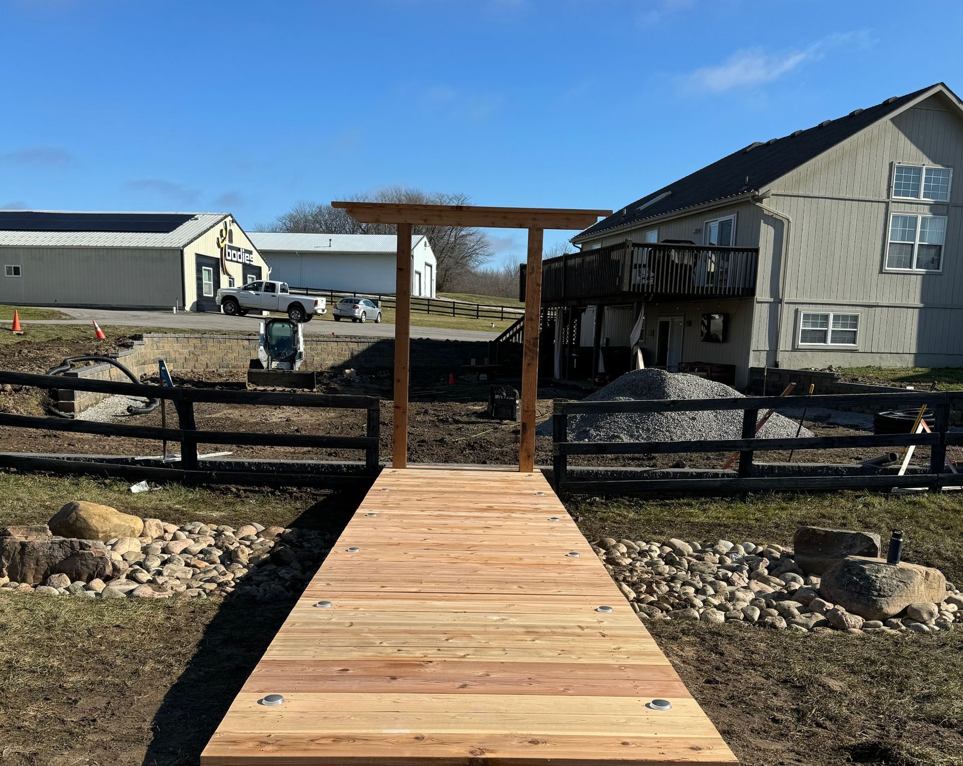 Wooden walkway leading to a fenced ranch with a gray house and barns under a clear sky