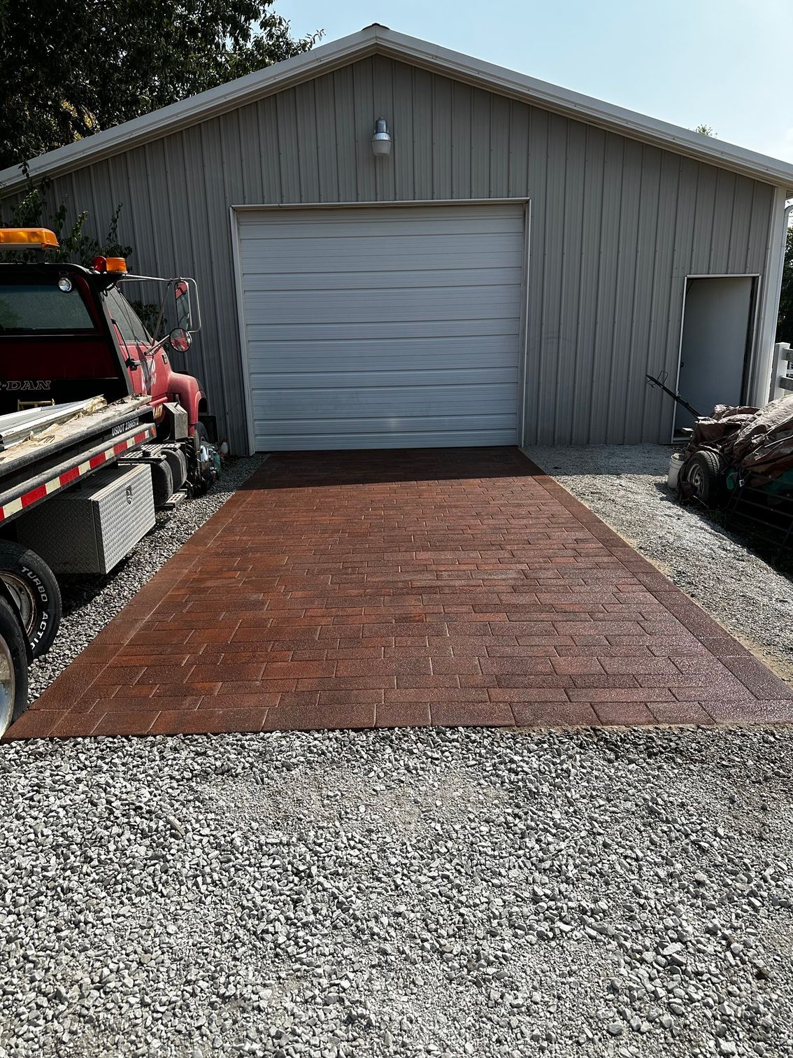 Gray garage with closed door, red concrete driveway, and gravel foreground; truck partly visible on the left