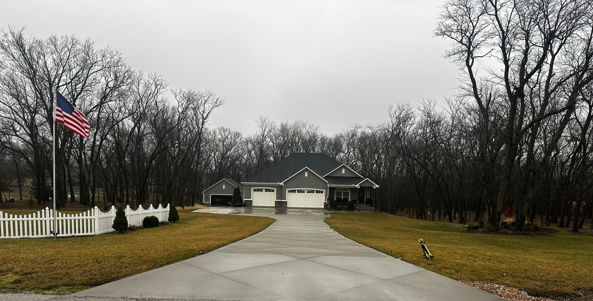 Curved driveway leading to a house on a rainy day, with an American flag and white fence on the left.